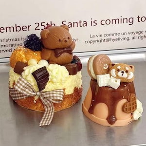 Peut inclure: Deux gâteaux décoratifs. L'un est un gâteau rond avec un ours en peluche sur le dessus, décoré de chocolat, de baies et d'un ruban à carreaux. L'autre est un petit gâteau avec un panda roux et un macaron sur le dessus, avec un filet de chocolat.