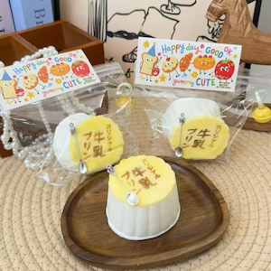 May include: A close-up of three desserts, two in clear plastic bags and one on a wooden plate. The desserts have a white base and a yellow top with Japanese text. Each dessert has a small spoon. The bags have a sticker that says "Happy day GOOD OK" and "yummy CUTE".