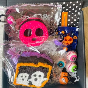 Op de afbeelding: Een Halloween-thema geschenkdoos gevuld met diverse griezelige items. Inclusief een roze schedel, een stuk cake met geesten, oogbal snoepjes en een pompoen beeldje. De doos heeft een stippen binnenkant en een "Happy Halloween" label.