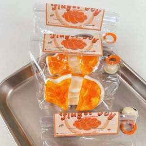 May include: Three clear plastic bags containing orange and white fidget toys shaped like fish. Each bag has a label with the words "Fidget Toy" and a graphic of orange shapes. The bags are on a silver tray.