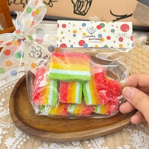 Può includere: Un sacchetto trasparente pieno di caramelle gommose multicolore a strati arcobaleno. Il sacchetto è legato con un nastro a pois e ha un'etichetta con la scritta "Happy Everyday". Le caramelle sono disposte su un piatto di legno.