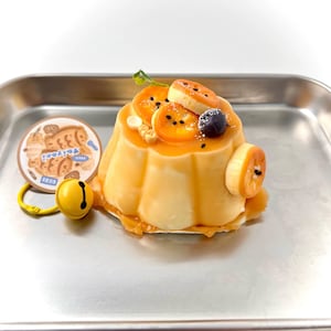 May include: A dessert resembling a pudding, topped with fruit slices, a blueberry, and a drizzle of sauce. A small, round charm with a bear design and a yellow bell are next to the dessert, all on a silver tray.