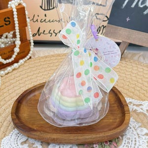 May include: A small, rainbow-colored cake, wrapped in clear cellophane and tied with a polka-dot ribbon, sits on a wooden plate. A small tag with the words "Sweet Moments, Beautiful Memories" is attached. The cake is a pastel rainbow of colors.