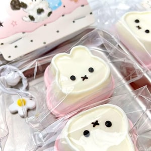 May include: Close-up of several bunny-shaped treats with white and pink coloring, black facial details, and wrapped in clear plastic. A small keychain with a white ball and a yellow flower is visible in the foreground. A pink and white box with a cloud design is in the background.