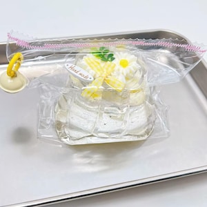 May include: A small, white cake with a floral design, wrapped in clear plastic. The cake is decorated with white frosting, yellow ribbon, and daisy-shaped decorations. The text "Hand made" is visible on a small tag.