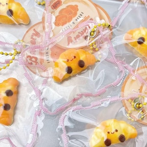 May include: Overhead view of cartoon croissants with smiling faces and chocolate chip eyes, packaged in clear plastic. Pink ribbon and yellow beaded accents are also visible. Bakery-themed paper circles with text are also present.
