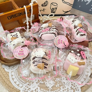 Peut inclure: Assortiment de douceurs décoratives emballées, dont des friandises en forme d'ours et de beignets, présentées sur une nappe en dentelle. Les sucreries sont principalement roses, blanches et marron, avec des éléments décoratifs. Une boîte en bois avec le mot "ORIGINALS" est visible.