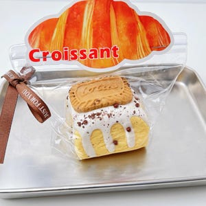 Op de afbeelding: Een gebakje in een doorzichtige zak, gegarneerd met een koekje en bedekt met witte glazuur. De zak is verzegeld met een bruin lint met de tekst "JUST FOR YOU". De zak is gelabeld "Croissant" in rode letters.