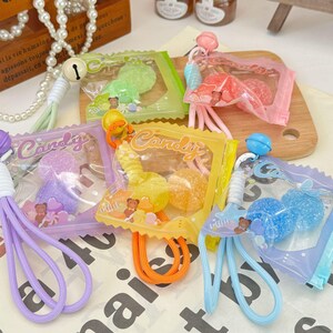 Può includere: Una collezione di portachiavi con caramelle colorate. Ogni portachiavi presenta un piccolo sacchetto di caramelle gommose in vari colori, tra cui verde, rosa, giallo, viola e blu. I sacchetti sono attaccati a un anello di cordino colorato.