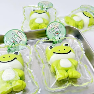 May include: Several green frog-shaped treats with white bellies are packaged in clear plastic. Each treat has a smiling face and is topped with a round label that reads "Ice Cream". The treats are arranged on a silver tray.