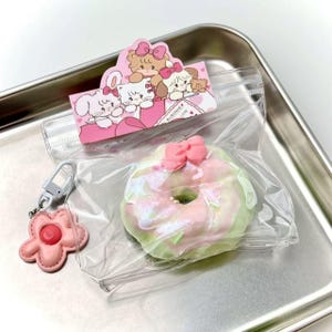 May include: A pink and green iridescent donut with a pink bow, packaged in a clear plastic bag. A pink flower keychain and a pink card with cartoon animals are also in the image, all on a silver tray.