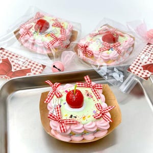 Può includere: Torte quadrate decorative con glassa rosa e bianca, guarnite con una ciliegia rossa e una piccola forchetta. Ogni torta è adornata con fiocchi a scacchi rossi e bianchi. Le torte sono confezionate in plastica trasparente e presentate su un vassoio di metallo.