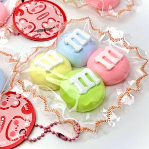 Può includere: Una collezione di caramelle rotonde e colorate in tonalità pastello di rosa, blu, giallo e verde, ognuna sormontata da un design a "M" bianco. Le caramelle sono confezionate in plastica trasparente con un bordo decorativo e un'etichetta rosa con la parola "Rabbit".
