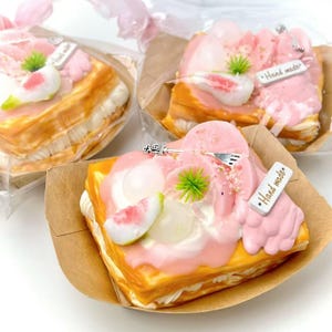 Könnte beinhalten: Drei dekorative, mehrschichtige Gebäckstücke mit rosa Glasur, weißer Sahne und Fruchtscheiben. Jedes Gebäck befindet sich in einem braunen Papierbehälter und ist in Klarsichtfolie verpackt. Kleine Metallanhänger mit der Aufschrift "Hand made".