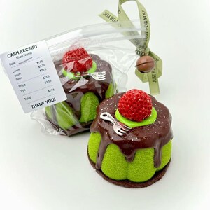 May include: Two green, cake-shaped soaps with chocolate frosting and a red strawberry on top. Each soap has a silver fork charm. One is in a clear bag with a receipt that says "THANK YOU!" and a green ribbon.