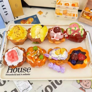 Puede incluir: Una bandeja de miniaturas de pasteles y postres coloridos. La bandeja incluye varias tartas con coberturas de frutas, una bola de helado y un caramelo. La bandeja tiene la palabra "House" y "Household and A esthetic" impresa.