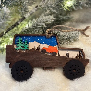 May include: A wooden Christmas ornament shaped like a Jeep driving through a snowy mountain scene. The ornament is painted with blue sky, white snow, and green trees. The Jeep is orange and has black wheels.
