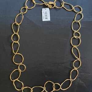 Pode incluir: Um colar dourado com uma série de elos ovais e circulares interligados. O colar tem um fecho de lagosta e uma pequena etiqueta anexada. Os elos variam em tamanho e forma, criando um design texturizado. A joia é exibida em um fundo escuro.