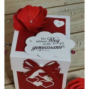 Könnte beinhalten: Eine weiße Box mit rotem Deckel, verziert mit einer roten Papierblume und einer weißen Wolke mit dem Text "Der schönste Weg ist der gemeinsame" in Schwarz. Die Box hat ein rotes Band und ein rotes Herzdesign auf der Vorderseite.