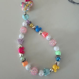Puede incluir: Un llavero de cuentas de colores con un cierre dorado. El llavero presenta una variedad de cuentas, incluyendo cuentas rosas, azules y amarillas con forma de Mickey Mouse, cuentas en forma de corazón y cuentas brillantes. La cuenta superior es una cabeza de Mickey Mouse arcoíris con un lazo rosa.