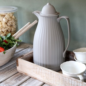 May include: A grey ceramic thermal carafe with a ribbed design sits on a wooden tray with white ceramic teacups.