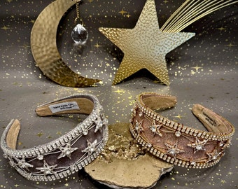 May include: Two embellished headbands with star and rhinestone accents. One is silver and the other is rose gold. The headbands are displayed on a gold-colored surface with a celestial background featuring a gold crescent moon and star.