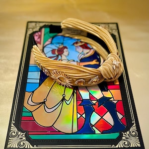 May include: A gold satin headband with a knotted bow and gold embellishments. The headband is on top of a black book with a stained glass image of Belle and the Beast from Beauty and the Beast.