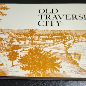 May include: A vintage book titled "OLD TRAVERSE CITY" with an illustration of a townscape in shades of orange and white. The cover features the text "From Original Photographs". The book is lying on a dark surface.