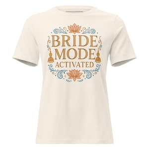 May include: Cream-colored t-shirt with a circular design featuring the words "BRIDE MODE ACTIVATED" in gold lettering. The design includes lotus flowers, swirls, and decorative elements, creating a festive look. A casual tee for a bride.