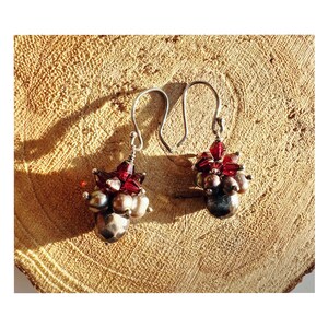 Könnte beinhalten: Silberne Ohrringe mit einer Ansammlung dunkelroter und grauer Perlen. Die Ohrringe haben einen Hakenverschluss und werden auf einer strukturierten, hellbraunen Oberfläche präsentiert. Ein Schmuckstück.