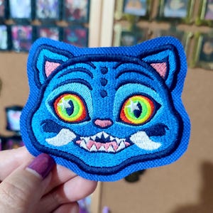 Puede incluir: Parche bordado de gato azul con una amplia sonrisa, dientes afilados y ojos grandes y coloridos. Los ojos del gato son amarillos y verdes, con detalles en rojo y naranja. El parche tiene un borde azul.
