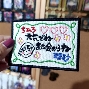 Puede incluir: Parche bordado con texto japon&eacute;s, una cara de dibujos animados sonriente, corazones y flores. El parche tiene un fondo blanco con un borde negro y costuras verdes. El texto es en negro y azul.