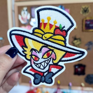 May include: Embroidered patch featuring a cartoon character with a top hat, crown, and bow tie. The character has a yellow face, red eyes, and a wide, toothy grin. The patch has a white border and is held by a hand.