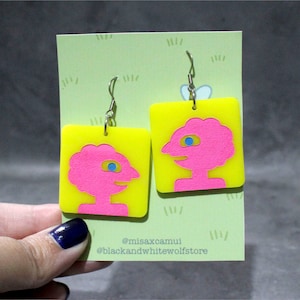 Puede incluir: Un par de pendientes cuadrados de color amarillo neón con un recorte rosa de una cara con un ojo azul. Los pendientes cuelgan de ganchos plateados. Se exhiben en una tarjeta verde claro con diseños de hierba y el texto "@misaxcamui" y "@blackandwhitewolfstore".