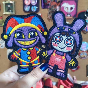 May include: Two embroidered patches featuring cartoon characters. The left patch shows a purple jester with a red and blue hat and suit. The right patch depicts a character in a purple bunny hood, wearing red overalls with star-patterned eyes.