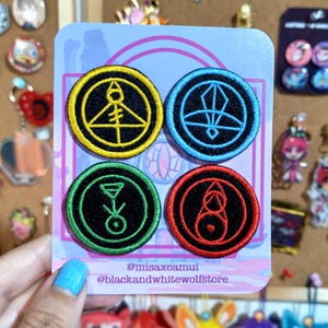May include: Four embroidered patches with circular designs in yellow, blue, green, and red. Each patch features a black border and a unique symbol. The patches are displayed on a light blue card with text that reads "@misaxcamui @blackandwhitewolfstore."