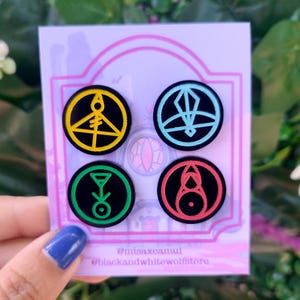 Peut inclure: Quatre épingles en émail avec différents symboles en jaune, bleu, vert et rouge sur fond noir. Les épingles sont présentées sur une carte avec le texte "@misaxeamul @blackandwhitewolfstore".