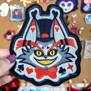 Puede incluir: Parche bordado con un personaje de gato de dibujos animados con sombrero de copa y pajarita. El parche tiene una combinación de colores gris, blanco, rojo y negro. El gato tiene ojos amarillos y una amplia sonrisa. Símbolos de juego en la parte inferior.