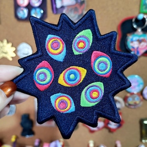 May include: A dark blue star-shaped embroidered patch featuring seven colorful eye-like motifs. Each eye design consists of concentric circles in vibrant shades of pink, yellow, green, and light blue, stitched onto a dark navy fabric base.