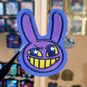 Può includere: Una toppa ricamata viola a forma di testa di coniglio con un sorriso sinistro. Il coniglio ha denti gialli, occhi a forma di stella e un contorno blu scuro. La toppa è tenuta da una pinzetta.