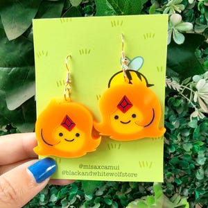 Puede incluir: Un par de pendientes de personaje de dibujos animados de color naranja y amarillo con ganchos dorados. Los pendientes presentan una cara sonriente con una gema roja y detalles negros. Los pendientes están sobre una tarjeta verde con el texto "@misaxcamui" y "@blackandwhitewolfstore".