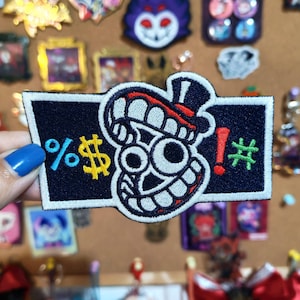 May include: Embroidered patch with a cartoon face design. The patch features a black background with white, red, yellow, and green accents. The design includes the symbols %, $, !, and #.