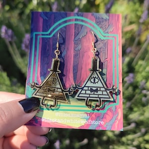 Puede incluir: Pendientes de color dorado con un personaje de dibujos animados con cabeza triangular, un ojo y una pajarita. El personaje está sobre un fondo rosa y morado con el texto "@misaxcamui @blackandwhitewolfstore".