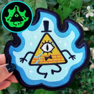 Puede incluir: Parche bordado con un triángulo amarillo con un ojo, sombrero de copa y pajarita, rodeado de llamas azules. El parche tiene un borde negro y una versión verde brillante más pequeña en la esquina.