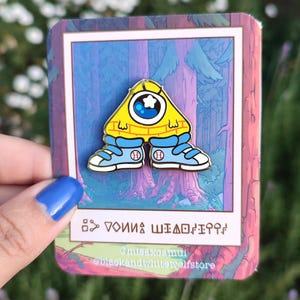 Puede incluir: Un pin de esmalte amarillo y azul con un personaje de dibujos animados con un solo ojo y una gorra de béisbol. El personaje lleva zapatillas azules con cordones blancos y una camisa a rayas amarilla y azul. El pin está en una tarjeta blanca con un fondo rosa y azul. El texto en la tarjeta dice "DON'T BE A SHADOW" y "@misaxcami @blackandwhitewolfstore".