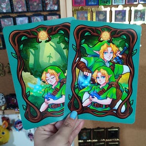 May include: Two rectangular art prints depicting a character from a video game. The character has blonde hair, green clothing, and holds a blue object. The prints are framed by a brown, leafy border with a green backdrop.