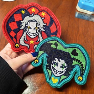 May include: Two embroidered patches featuring jester characters. The top patch is red and navy blue with a white-haired jester, yellow stars, and a red-and-navy checkered pattern. The bottom patch is teal and black with a dark-haired jester and hearts.
