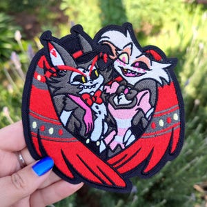 Könnte beinhalten: Gestickter Aufnäher mit zwei Comicfiguren, eine mit roten Hörnern und einem Zylinder, die andere mit weißem Fell und einer rosa Schleife, beide mit lächelnden Gesichtern. Die Figuren sind von roten Flügeln mit schwarzen und weißen Akzenten umgeben.