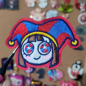 Può includere: Toppa ricamata raffigurante un giullare dei cartoni animati con un cappello rosso e blu, capelli castani e un viso sorridente. Gli occhi del giullare hanno un disegno a stella. La toppa è bordata di rosso e ha una fascia gialla intorno alla testa.
