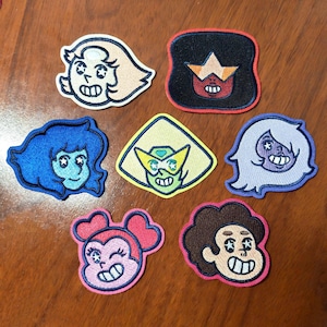 May include: A collection of seven embroidered patches featuring cartoon character faces. The designs include a light-haired figure, a dark-haired figure with a star, a blue-haired figure, a green-haired figure with goggles, a purple-haired figure, a pink-haired figure, and a brown-haired figure.